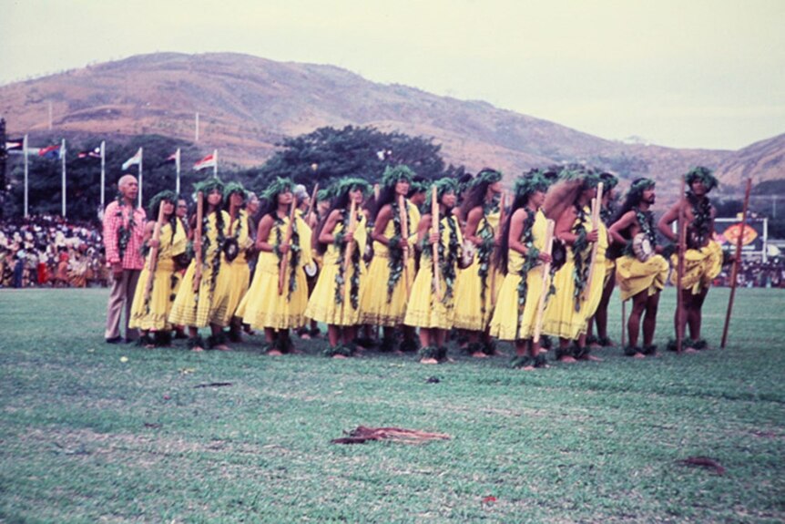 Group of people in Hawaiian cultural dress stand on a field outdoors. Mountains and flags in background. 