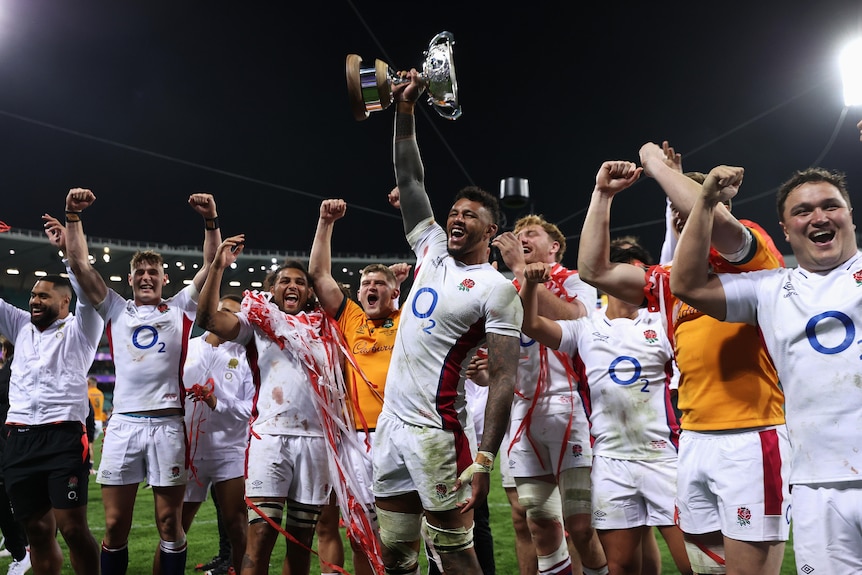 The English men's rugby union team raise their arms in triumph as the captain lifts a cup high.