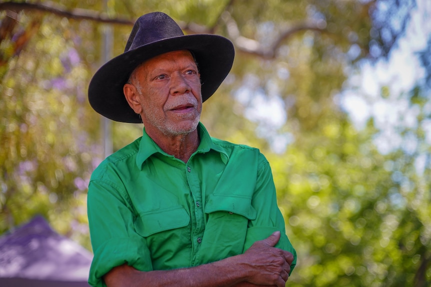 An Indigenous man wearing an akubra standing in a park, with a neutral expression.