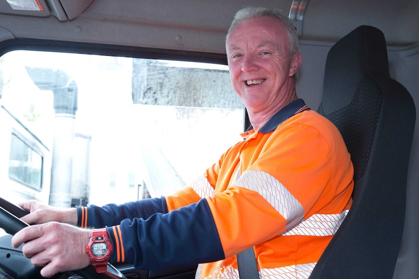John Pavitt smiling in orange hi-vis vest behind the wheel of his truck. He has short grey hair.