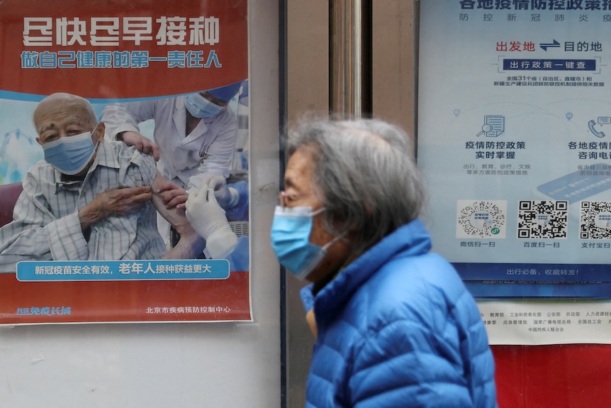An elderly woman wearing a face mask walks past a poster showing an elderly man getting a needle