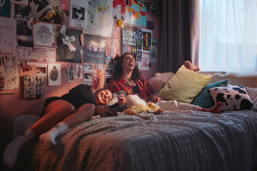 Two young women lie around on top of a bed, laughing
