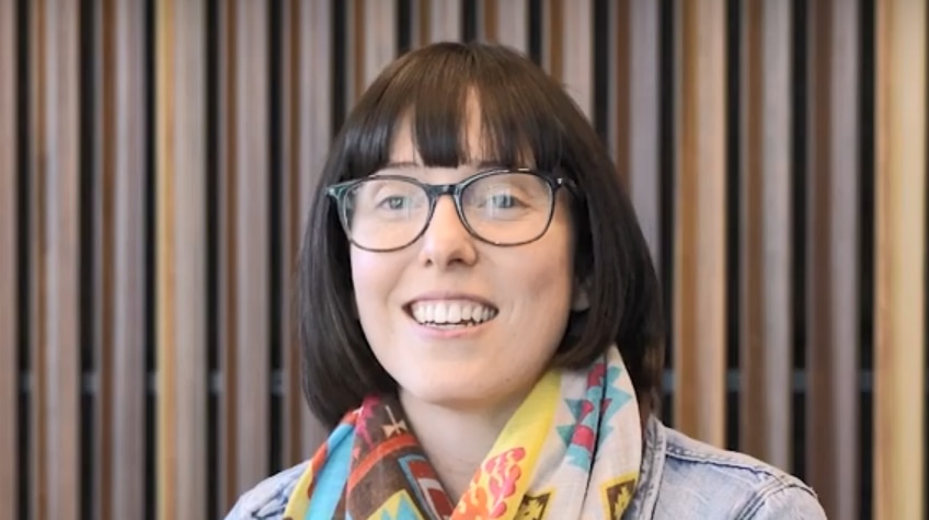 Woman wearing reading glasses with short hair sits facing camera and smiles