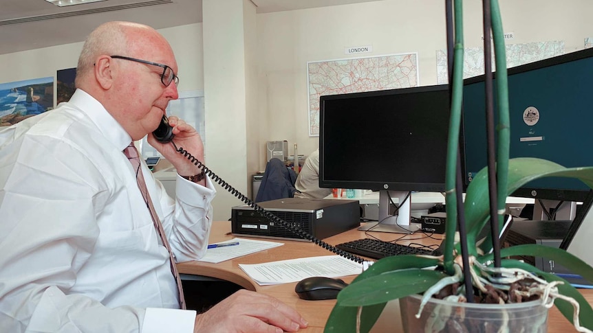 George Brandis speaks on a phone while sitting at a desk.