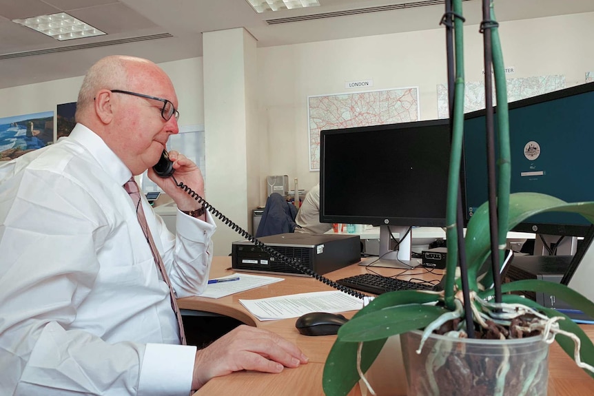 George Brandis speaks on a phone while sitting at a desk.