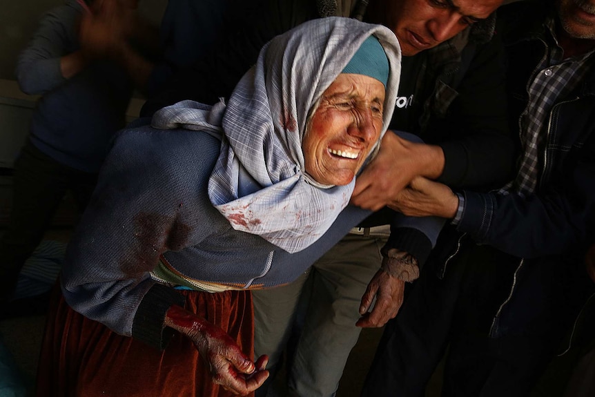 An older woman covered in blood cries as people try to comfort her.