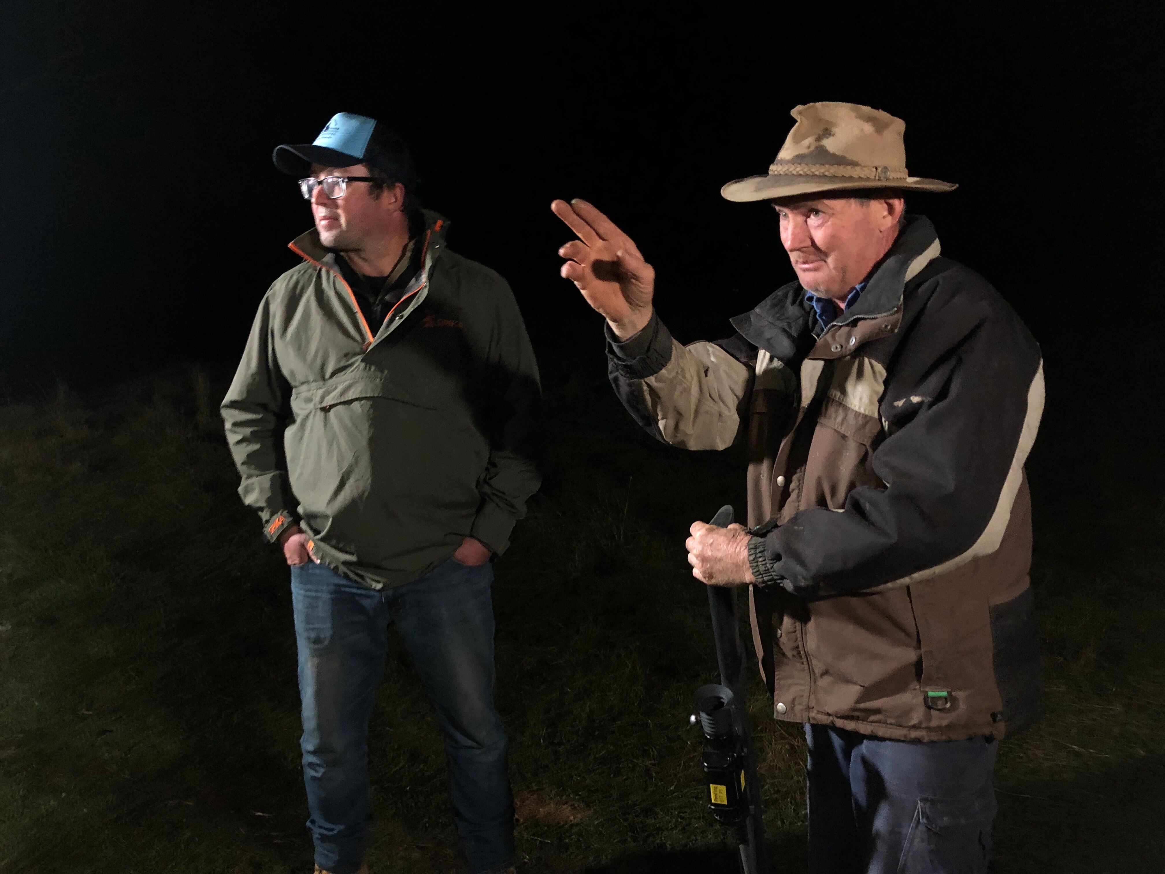 Two men stand side by side at night, both dressed in warm outdoor wear. One gestures a direction.