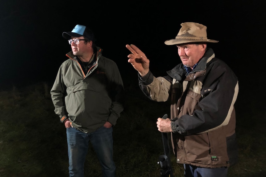 Two men stand side by side at night, both dressed in warm outdoor wear. One gestures a direction.