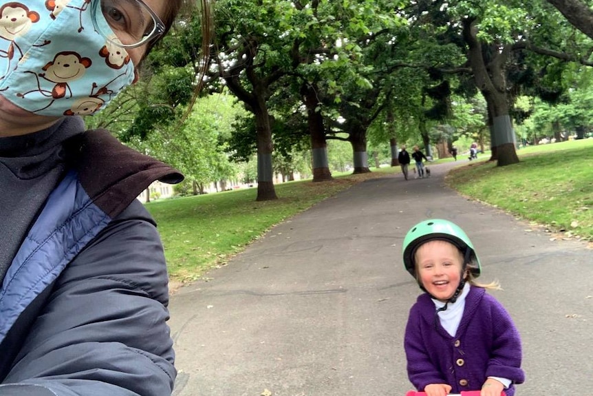 Woman wearing a mask takes a self with a child on a scooter.