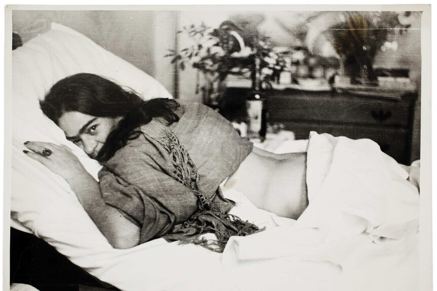 Frida Kahlo lies face down on a bed, her head turned to look at the camera.