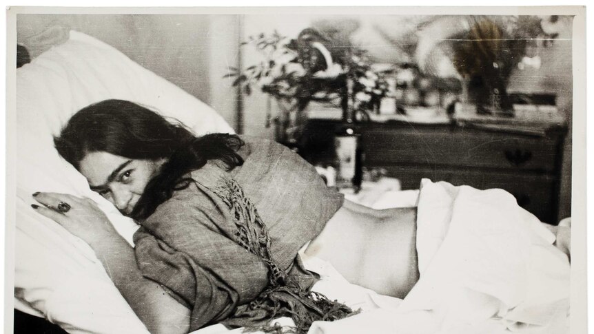 Frida Kahlo lies face down on a bed, her head turned to look at the camera.