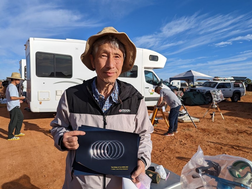 A middle-aged man wearing a hat stands at a campsite holding a photo of a previous solar eclipse.