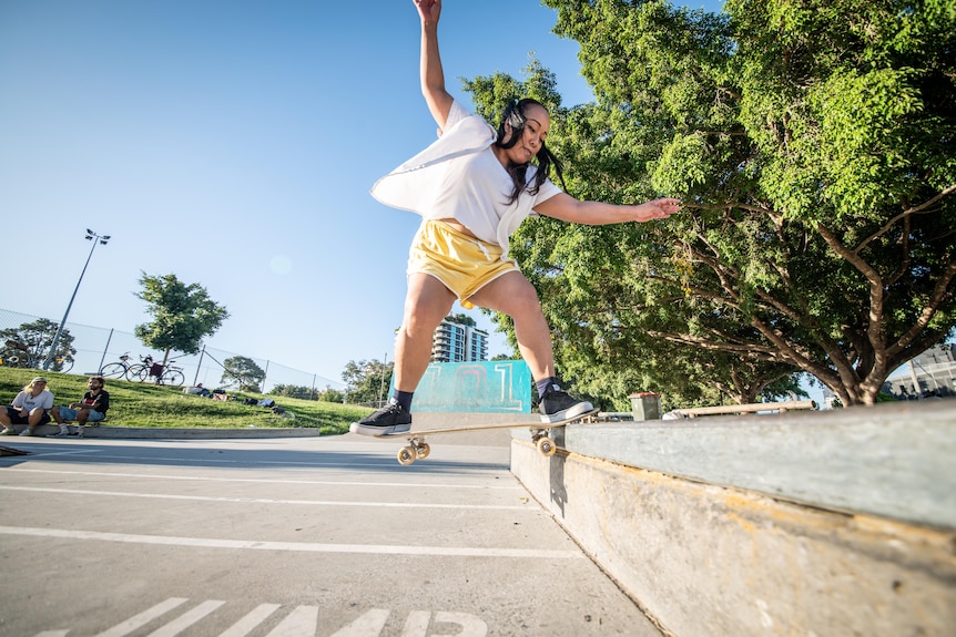 A woman wearing a white-t-shirt and yellow shorts is skating, with her board perched on the edge of a ledge as she balances