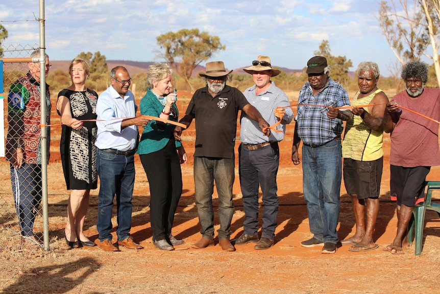 Nine people stand in a row as they cut an orange ribbon with scissors, against a backdrop of red dirt and gum trees.