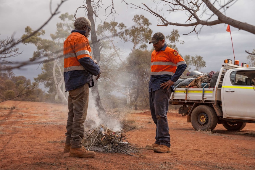 Two men, wearing high-viz shirts, stand around a campfire