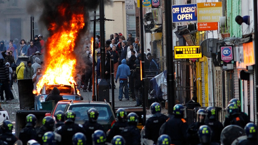 Police officers in riot gear block a road near a burning car on a street in Hackney, east London (Luke MacGregor)