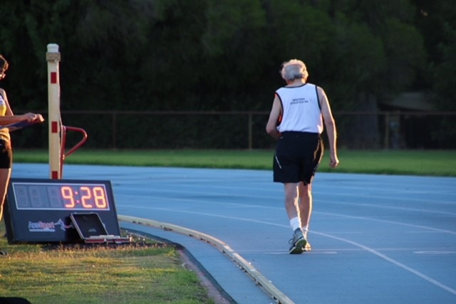 An elderly man on a blue running track next to a timing clock.
