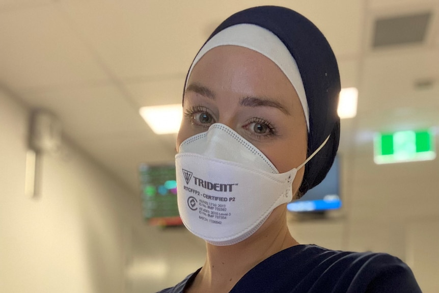 A woman in a nurses's uniform and a medical mask looks into the camera.