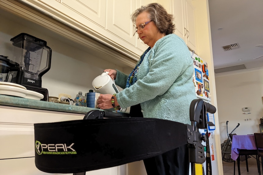 An older woman pouring a kettle in her kitchen with her walker in front of her.
