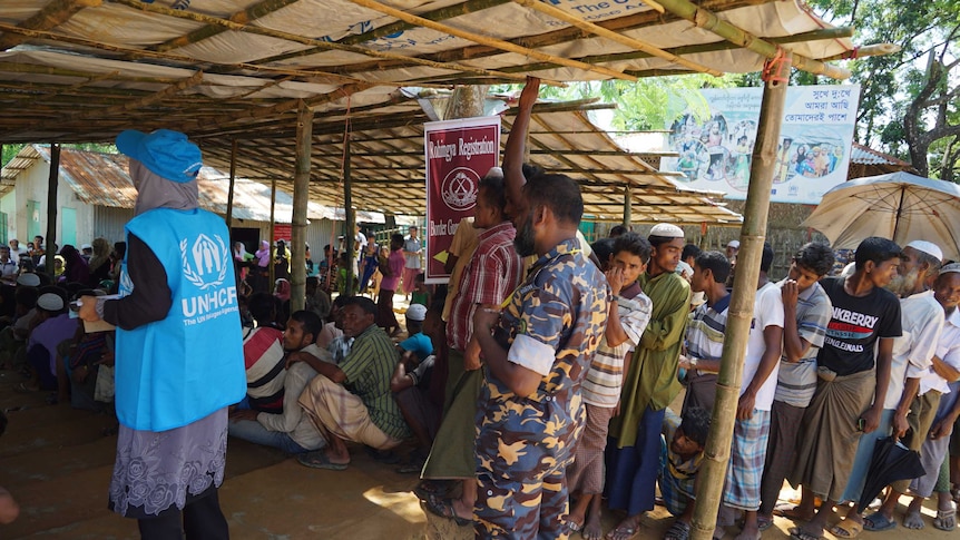 Men queue under a makeshift tent with a soldier and a person in a light-blue UN vest looking on.