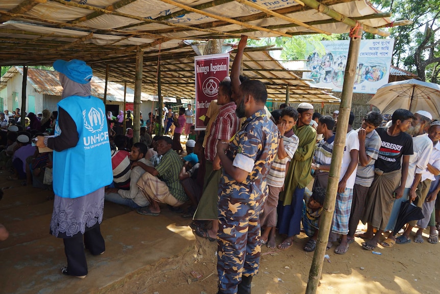 Men queue under a makeshift tent with a soldier and a person in a light-blue UN vest looking on.