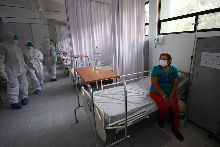An old woman in green wearing a face masks sits on a bed in a hospital ward, surrounded by staff.