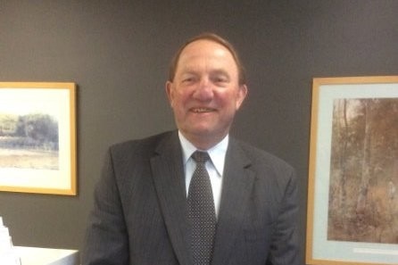 A man in a grey suit stands in an office, smiling while posing for a photo.
