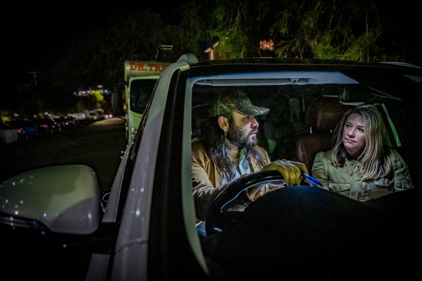 A man and a woman with pensive expressions sit in a car with the interior light on, in a dark street