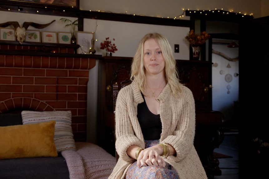 A woman with blond hair and cardigan sits in a chair 