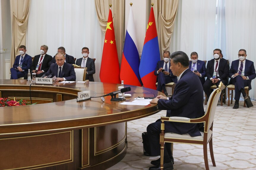 Putin and Xi sit at a massive, round table, as men in suits sit in chairs lining the walls.