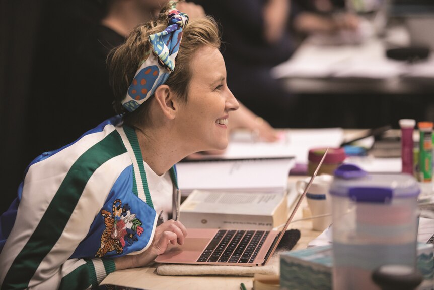 Kate Mulvany sitting at desk with laptop in front of her, wearing head scarf and bomber jacket.