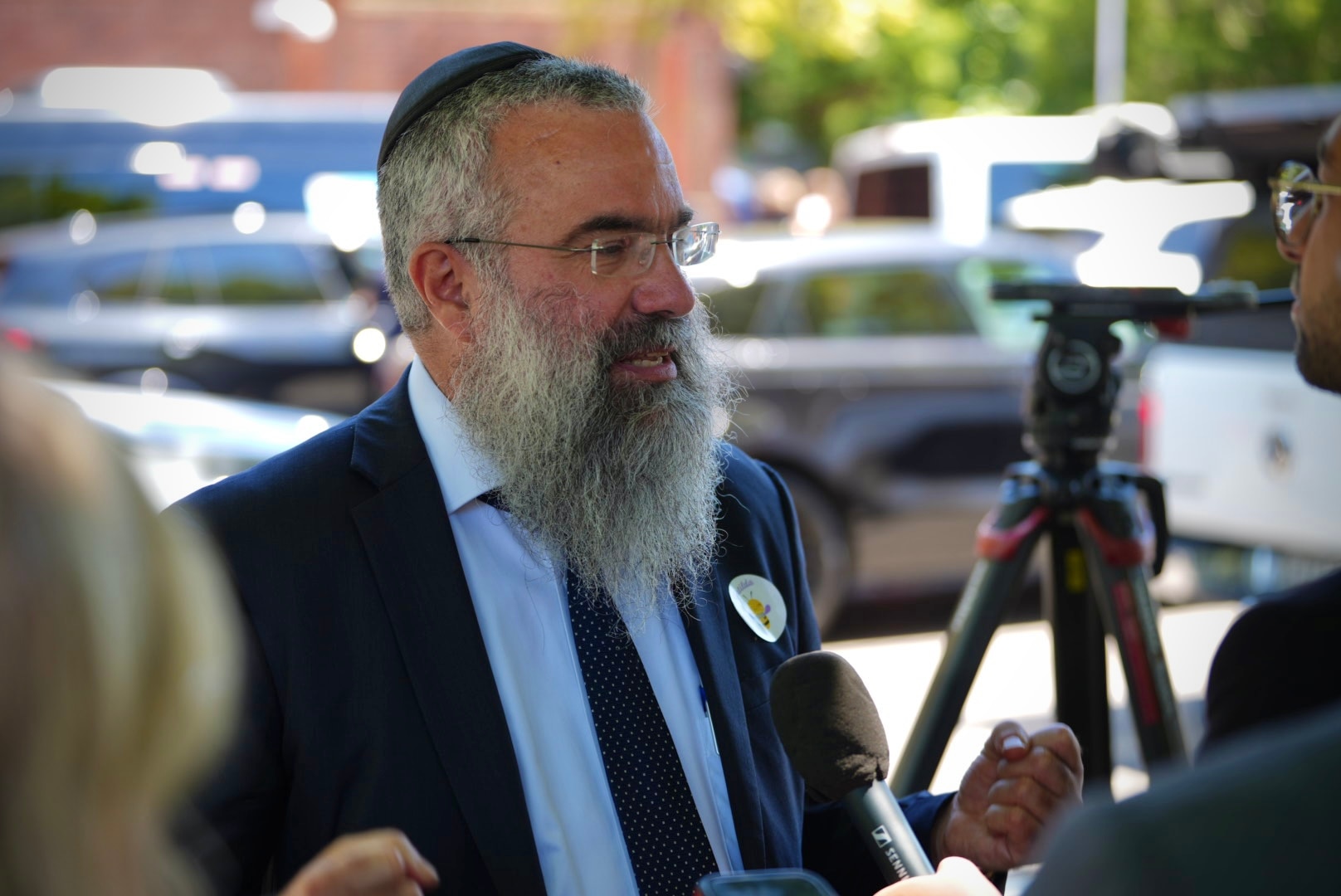 El rabino Dr. Dovid Slavin está de pie con traje, corbata y kipá con gafas y una pegatina de una abeja en su chaqueta.