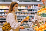 A woman wearing a face mask in a grocery store holds up a lemon.
