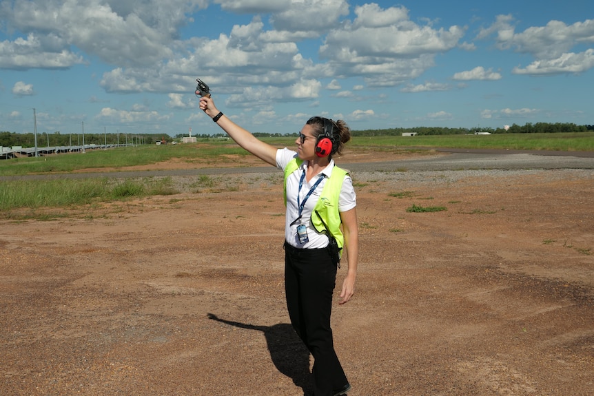 Woman in hi-viz vest shooting a firearm into the sky. Red dirt and green grass behind.