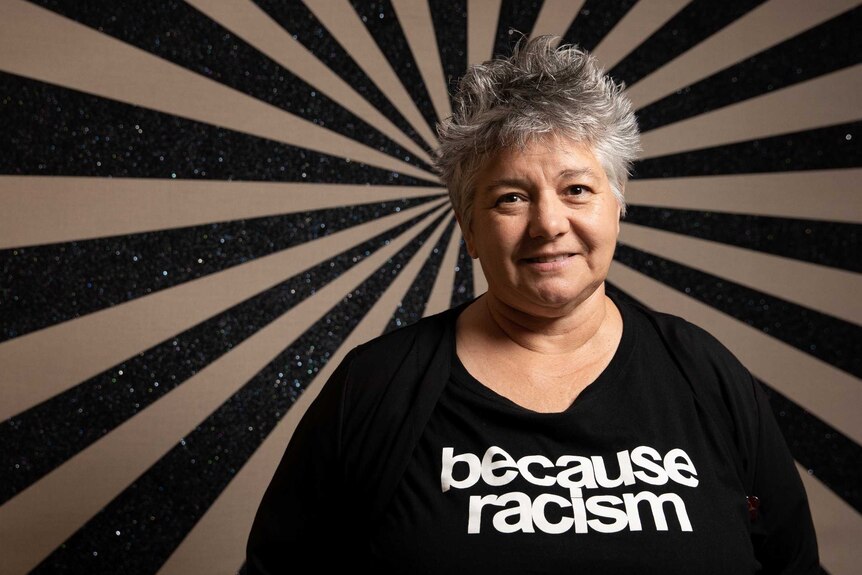 The novelist, wearing a black t-shirt that says "because racism", poses in front of a black and white painting.