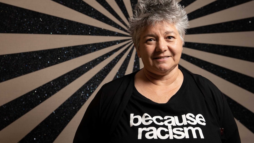 The novelist, wearing a black t-shirt that says "because racism", poses in front of a black and white painting.