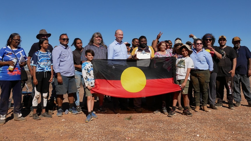 A man in a blue shirt stands in the middle of a crowd of Indigenous people, two children hold an Aboriginal flag.