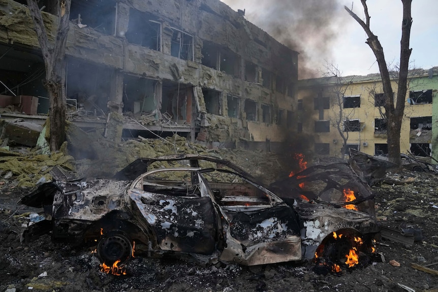 A burnt-out car is on fire outside of a badly damaged hospital building.