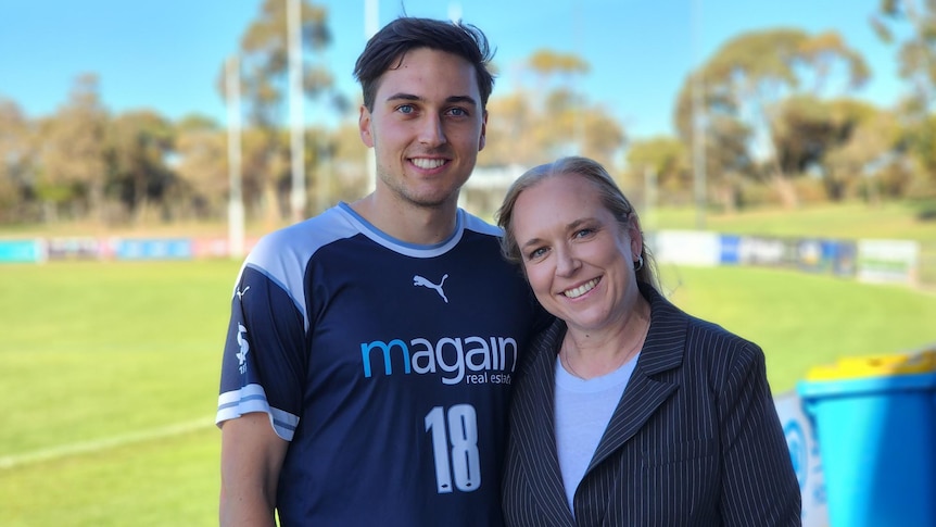 A smiling mother and son standing next to each other on a sunny day at a football field.