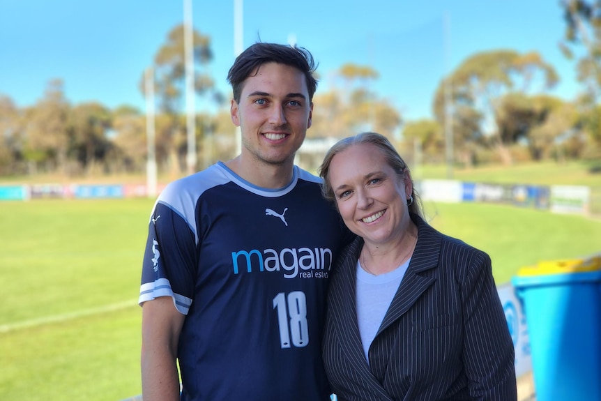 A smiling mother and son standing next to each other on a sunny day at a football field.