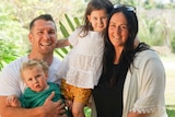 A woman and man smile together, holding their two young children. There are trees in the background.