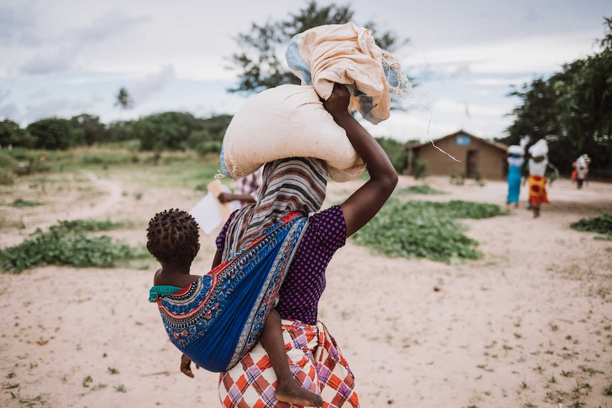 African woman carrying a sack of food on her head and a baby in a sling on her back, while walking towards a mud hut.