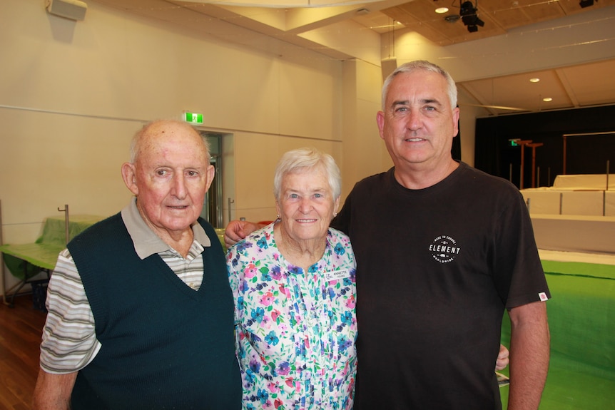 An older man and woman stand with their middle-aged son, all with their arms around each other.