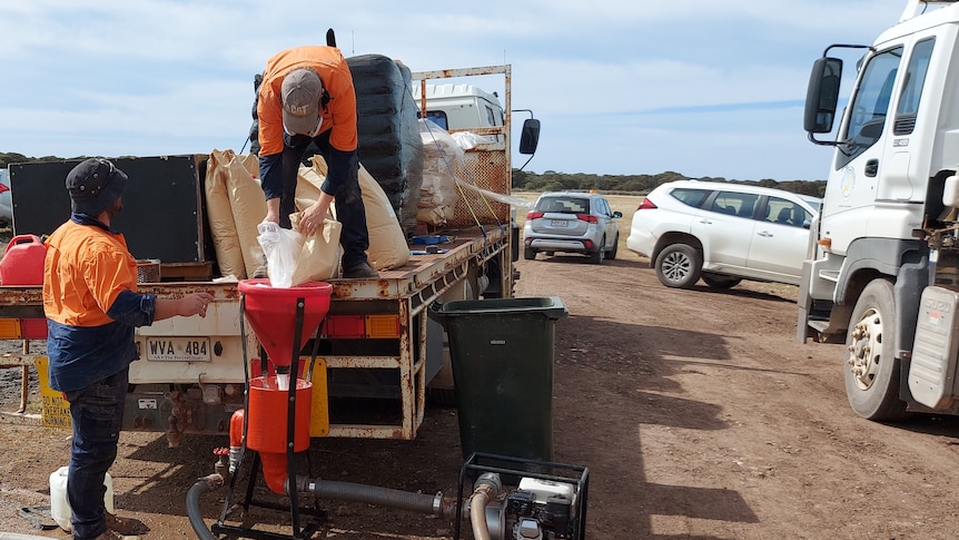 Workers in orange hi-vis stand on and around a truck pouring a powder into a red hopper.