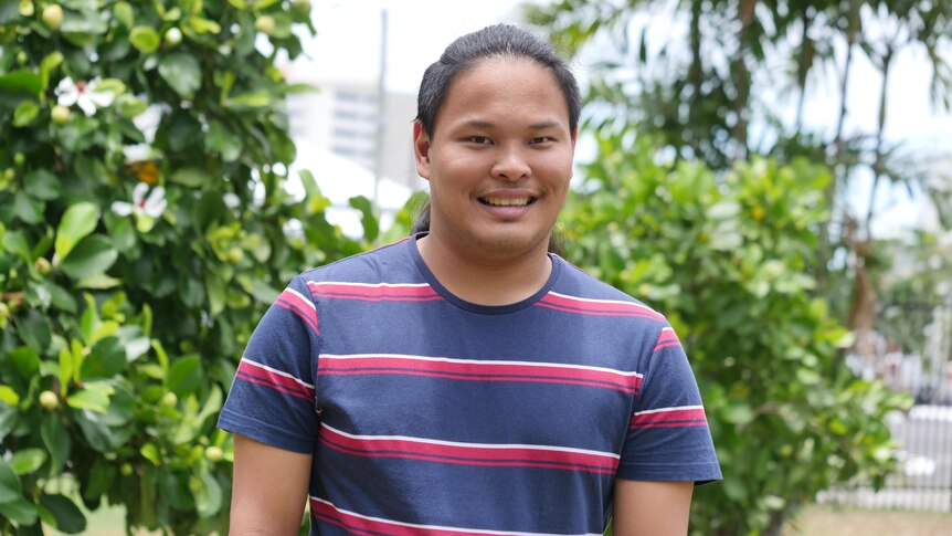 A young Asian man looks to the camera and smiles while wearing a navy t shirt with red and white stripes on it. 
