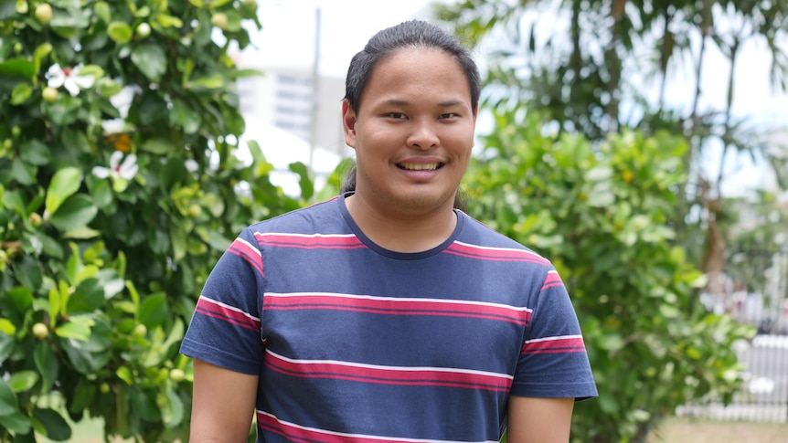 A young Asian man looks to the camera and smiles while wearing a navy t shirt with red and white stripes on it. 