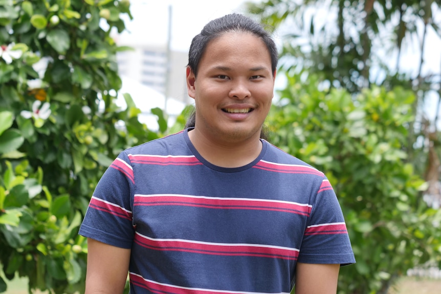 A young Asian man looks to the camera and smiles while wearing a navy t shirt with red and white stripes on it. 