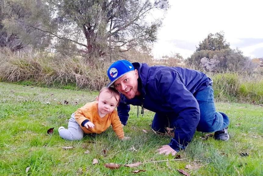 A man crouched down on the grass with a baby boy smiling at the camera