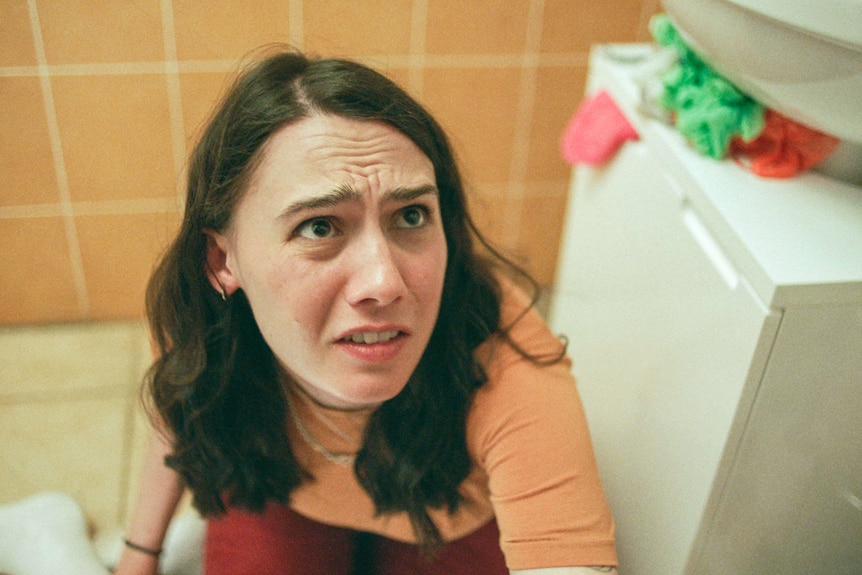 A young women with long brunette hair sits in a bathroom, looking worried