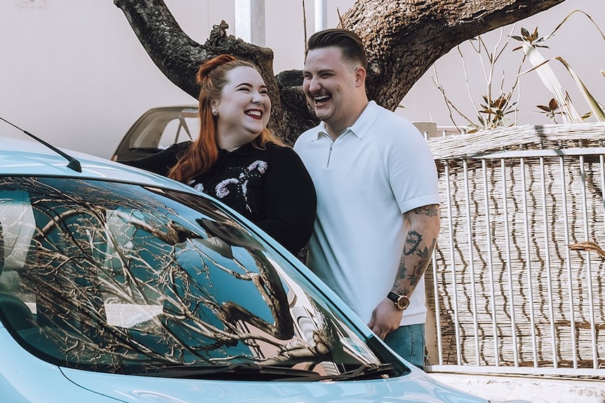 On a street, a woman and man stand close together behind a car, looking at each other and laughing happily.